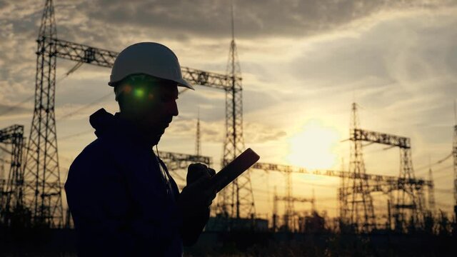 An electrical engineer wearing a helmet works with a tablet near high voltage electrical lines. An electrician works with the documents of an electrical substation. Energy business concept.