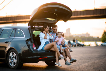 Young family with little daughter are resting while sitting on the trunk of the car