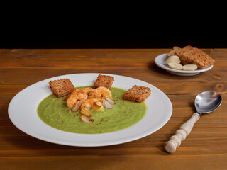 A plate of thick fresh green pea soup with prawns on a rustic wooden table with a garlic and croutons on the side.