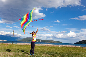 Kid playing with rainbow kite. Child girl has fun in summer holidays by sea landscape, mountains, blue sky. Happy childhood. Lifestyle moment. Outdoors activities with social distance in new normal.