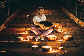 silhouette of a man playing handpan surrounded by Tibetan singing bowls and lighted candles sitting...