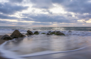 Amazing sea sunrise with slow shutter and waves flowing out around big rock at a sandy beach
