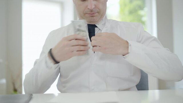Unrecognizable doctor looking around and hiding bribe in pocket. Corrupted medical employee having illegal income. Mid-adult Caucasian man earning money from bribery.