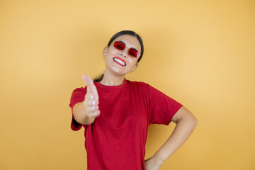 Young beautiful woman wearing red sunglasses over isolated yellow background smiling friendly offering handshake as greeting and welcoming.