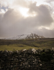 Dry stone wall with snow topped mountain in the background with sun breaking through the clouds