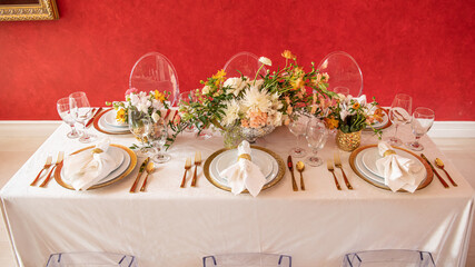 Elegant table setting with gold accents and red wall, clear chairs and flowers