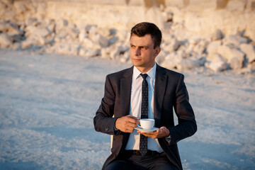 Young man in a classic jacket and tie is sitting on the chair and drinking coffee outdoors near a...