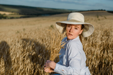 Naklejka na ściany i meble Woman in white shirt and hat is holding a bunch of wheat in the wheat field.