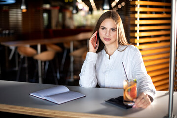 girl in a white shirt sits at a table and talks on the phone, next to a table is a drink and a notebook