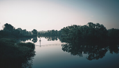 Dawn on the river. A fisherman is standing on the suspension bridge. Vegetation is reflected in calm water.