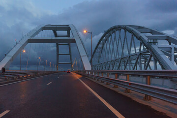 Cars drive under the arches of the Crimean bridge in the late evening. Lights are on