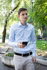 Portrait of young man in a blue shirt on the city with a phone in his hand with a trees on the background. The man in conversation, attentive listens. American or European appearance. Summer day.