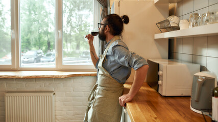 Single man in apron looking at the window, drinking wine from a glass while cooking in the kitchen