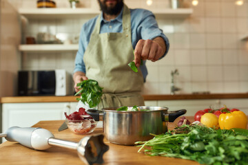 Cropped shot of man, chef cook adding basil leaf to the pot with chopped vegetables while preparing healthy meal, soup in the kitchen. Cooking at home, Italian cuisine concept