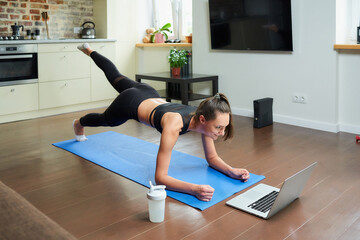A sporty girl in a workout tight suit is doing buttocks workout with an online video on a laptop. A female coach conducting a remote fitness class on the blue mat at home.