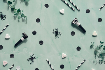 Halloween background on mint green stone. Flat lay with eucalyptus twigs and party decor - confetti, sugar hearts, whistles, drink straws and spiders.
