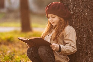 Cute little girl reading a book sitting under a tree in the park in autumn. Learning concept. Retro style. Children's fashion.
