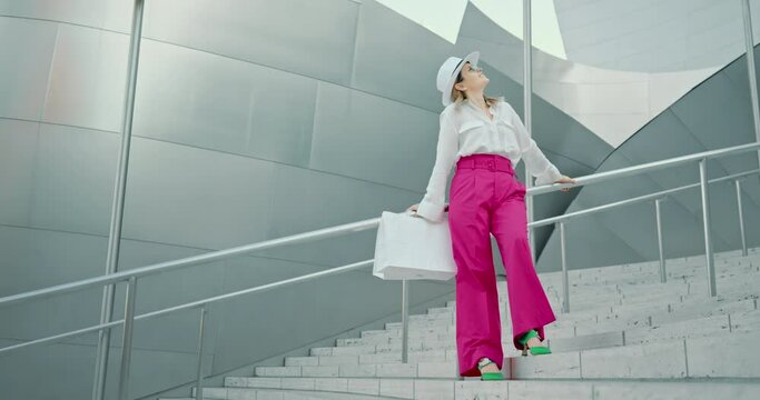 Woman in luxury outfit with pink bottom and white top and shopping bags is posing on stairs. Slow motion 4K fashion blogger posing for street style picture. Attractive young woman in urban downtown