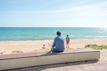 Back view of father relaxing reading book while children playing on sea beach, sunshine outdoors background. People dreaming romantic image.