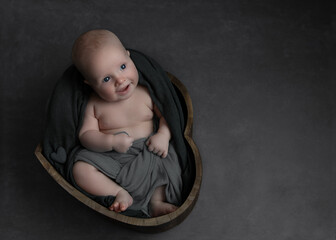 happy blue eyed baby boy smiling sitting in heart shaped wooden bowl draped in grey fabric with...