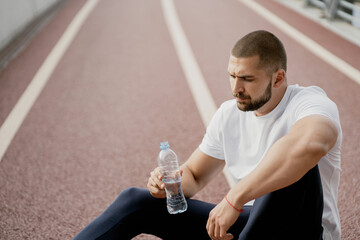 rest after sports training. a confident man is sitting on a treadmill, drinking clean water from a bottle. thinking about their health and plans for budushee. European appearance