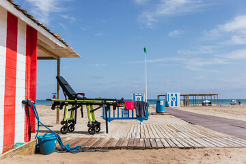 Stretcher on the beach next to the lifeguard hut with a wooden plank path on white sand and the mediterranean sea as a blurry background and blue sky