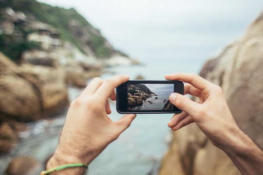 Close up of male hands holding a phone to take pictures of buildings next to the rocky coastline