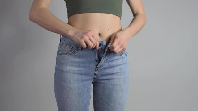 Slim woman is putting on a pair of jeans and buttoning zipper fastener then shows perfect waist after successful lost weight.