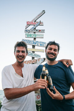 Two young men friends toasting beers next to a signpost with directions and distance to various countries