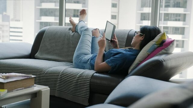 Happy female student reading ebook and lying on sofa. Young woman relaxing with book and man with smartphone. Hispanic people and lifestyle, leisure and relaxation at home