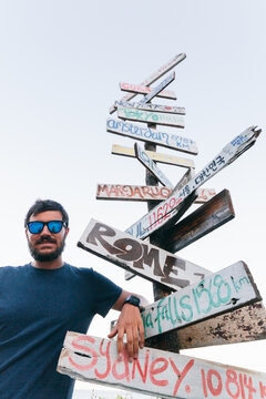 Young hipster man wearing sunglasses next to a signpost with directions and distance to various countries