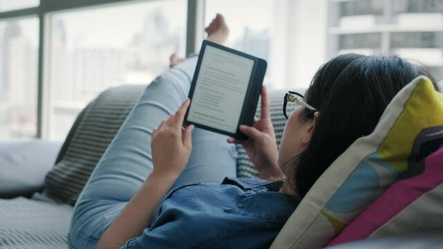 Happy female student reading ebook and lying on sofa. Young wife relaxing with book and husband with cell phone. Latino people and lifestyle, leisure and relax at home