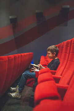 Young boy sitting alone in a movie theater waiting for a film to start and eating popcorn.
