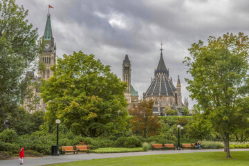 View of the Canadian Parliament Buildings Peace Tower and Parliamentary Library from Major's Hill Park, trees, park benches, sidewalk and lamp posts, lone girls in red coat walking alone on pathway un