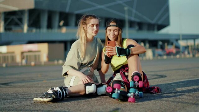 Stylish two young women sitting on ground with roller skates use phone smiling feel happy street in summertime legs rollerblading activity slow motion