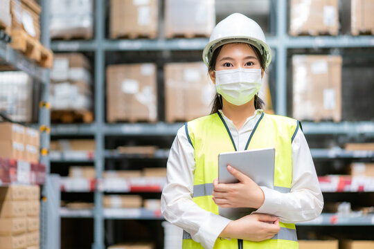 Young Asian Woman Auditor Or Trainee Staff Wears Mask Working During The COVID Pandemic In Store Warehouse Shipping Industrial. Looking Up And Checks The Number Of Items Store By Digital Tablet.