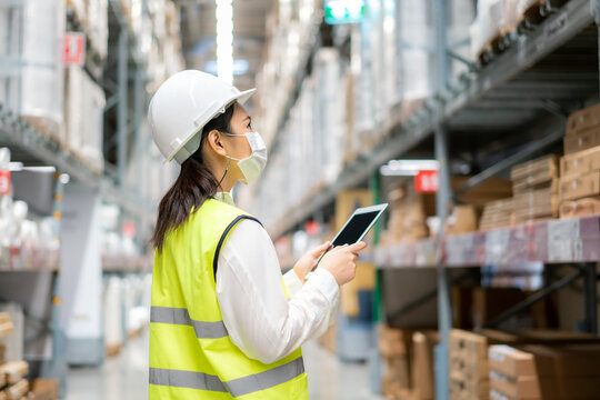 Young Asian Woman Auditor Or Trainee Staff Wears Mask Working During The COVID Pandemic In Store Warehouse Shipping Industrial. Looking Up And Checks The Number Of Items Store By Digital Tablet.