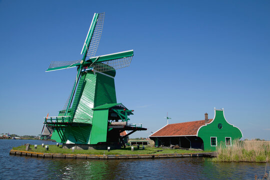 A classic windmill standing in the sun next to a green shed right along a dutch canal