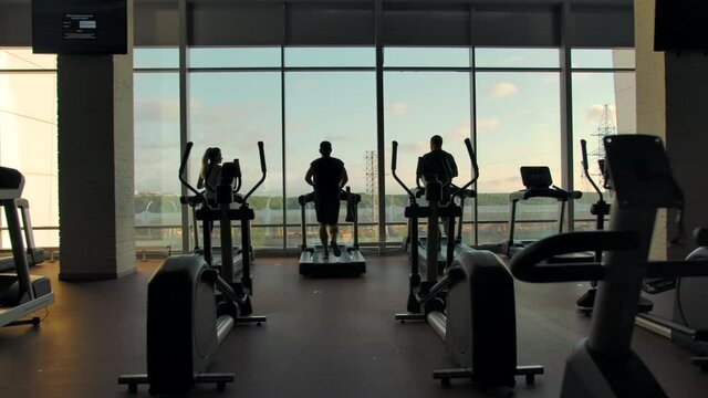 Silhouettes of three unrecognizable people running on treadmills in an empty gym against a large window on a sunny day. eft to right extreme slow motion shot.