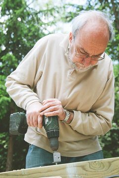 Old man with drill in outdoors making carpenter work at his garden.