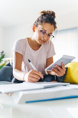 Front view on young female woman caucasian girl writing in notebook and reading on digital tablet while study at home - education and leisure concept