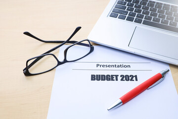 Text on budget 2021, a pen, eyeglasses, and laptop on wooden table 