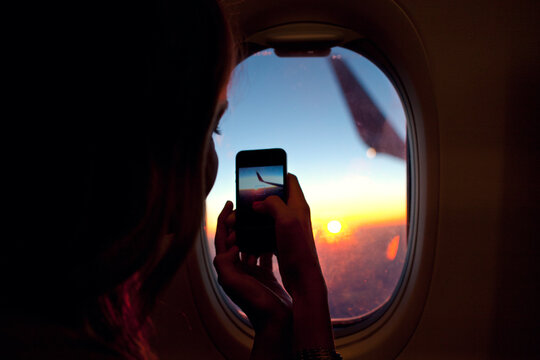 Young woman sitting in a window seat in an airplane and is using her phone to take a photo.