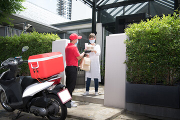 Asian delivery man wearing face mask and gloves in red uniform delivering bag of food and drink to...