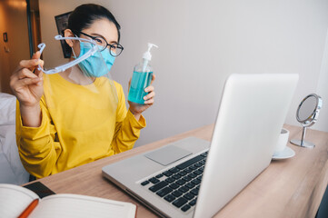 Asian woman holding gel sanitizer and plastic face shield while working online on laptop for advice her friends to using it when going outside during covid-19 pandemic.