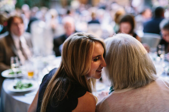 Young woman whispering secret message into ear of mother at table during dinner party