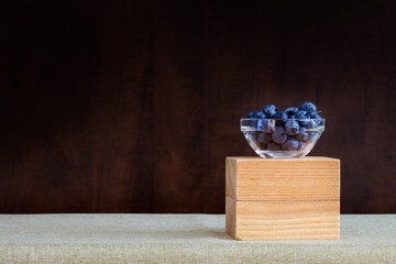 Fototapeta premium Small glass bowl of blueberries on a table with dark wood background behind; blueberries on a table covered with green table cloth
