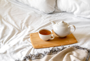 Obraz na płótnie Canvas Tray of tea on bed. White bedding sheets with blanket and pillow. Breakfast in bed. Warm and cosy scandinavian hygge concept - cup of tea.