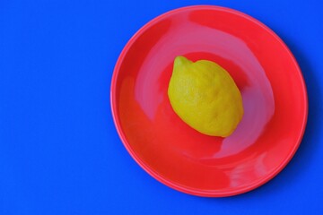 Lemon fruit in a minimal style. Bright yellow lemon in a red round plate on a  blue background.Graphic Fruits. Minimalist food. Citrus fruit concept.Copy space