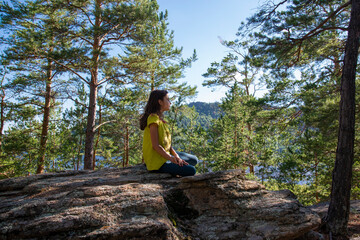 Naklejka na ściany i meble A girl with long hair is sitting on a rock in the forest against the background of a lake and pines.